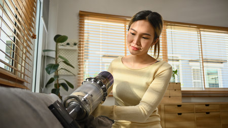 Beautiful female housewife vacuuming sofa with cordless handheld vacuum cleaner. Housework concept.の写真素材