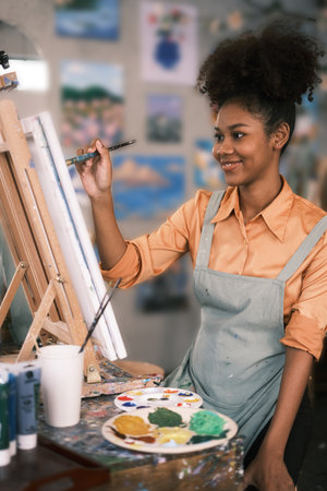 Cheerful young curly art school female student painting picture with brush on canvas in art studio.の写真素材
