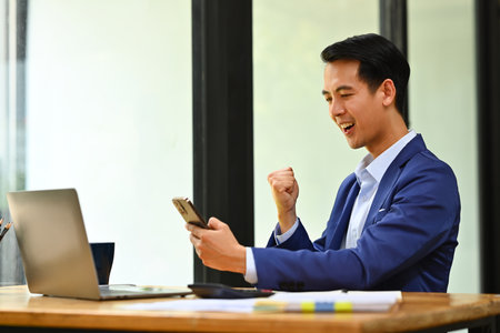 Surprised businessman looking at smart phone excited by good news online, celebrating success raising hands up and screaming.の写真素材