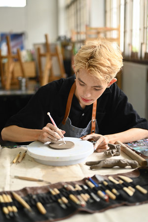 Focused man pottery artist decorating clay plate on table at ceramic workshop. Indoors lifestyle...