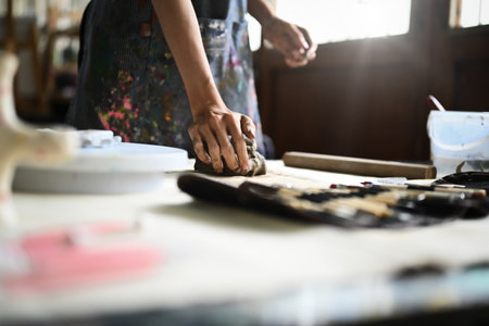 Craftswoman in apron kneading piece of raw clay with his hands on wooden table. Handicraft,...