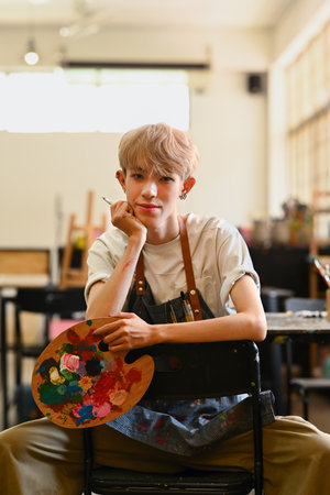 Portrait of young hipster male artist wearing apron holding palette and brush posing in art studio.の写真素材