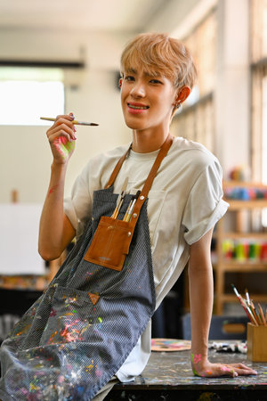 Cheerful Asian gay man wearing apron holding paintbrush sitting in art workshop. Hobby and leisure concept.の写真素材