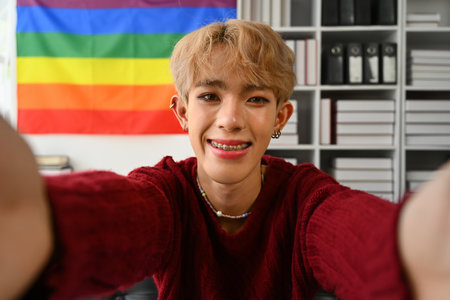 Gen z asian gay man holding camera taking selfie, shooting vlog or having virtual chat video call at home with rainbow flag on background.の写真素材