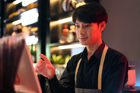 Handsome male waiter touching display of electronic cash register, entering customer order at bar or coffee shop.の写真素材