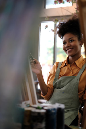 Delighted young woman with Afro haired wearing apron painting picture with brush on canvas in art studio.の写真素材