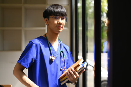 Portrait of medical student man wearing blue scrubs with stethoscope standing near window in sunny day in medical office.の写真素材