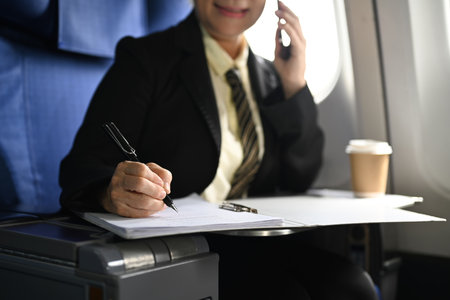 Smiling mature businesswoman having phone conversation and making important notes while sitting in airplane cabin.の写真素材