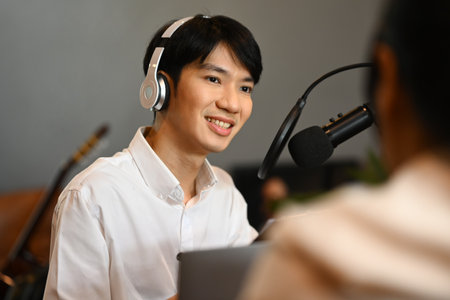 Friendly asian male broadcaster wearing headphone interviewing her guest at small home studio.の写真素材
