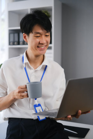 Asian male office worker holding cup of coffee and reading email on laptop, sitting at desk in...