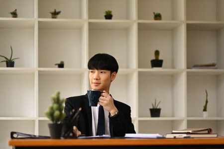Thoughtful Asian male entrepreneur sitting at office desk and looking away, thinking of future career vision.の写真素材
