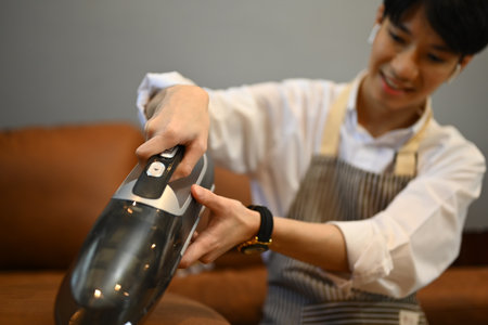 Happy young man cleaning sofa with cordless vacuum cleaner to removes germs and dirt. Housework concept.の写真素材