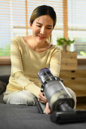 Smiling young woman vacuuming and cleaning the dust on couch with handheld cordless vacuum cleaner. Housework concept.の写真素材