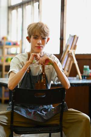 Young hipster queer artist holding palette and brush sitting in front of easel in bright studio. Art, education, creativity concept.の写真素材