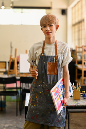 Young creative queer artist holding palette and brush standing in bright studio. Art, education, creativity and people concept.の写真素材