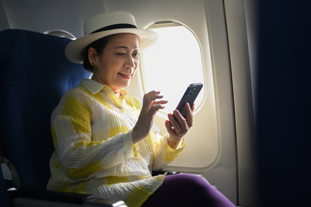 Cheerful senior woman tourist sitting in airplane cabin and using mobile phone waiting for airplane...