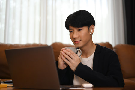 Pleasant man in wireless earphones and casual clothes drinking coffee and using laptop in living room..の写真素材