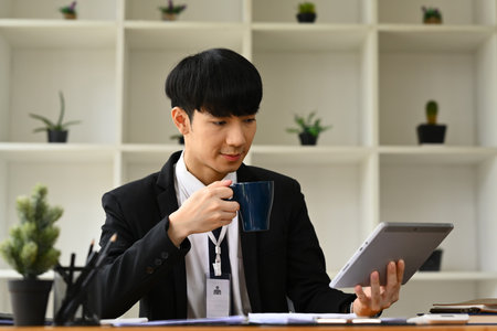 Male economist drinking coffee and using digital tablet, working with sales statistics document at office desk..の写真素材
