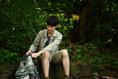 Man with backpack dressed in active trekking clothes sitting on dried tree trunk while exploring nature..の写真素材