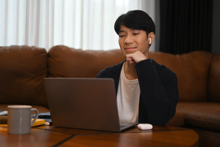Smiling man in wireless earphones and casual clothes browsing internet or working online on laptop at homeの写真素材