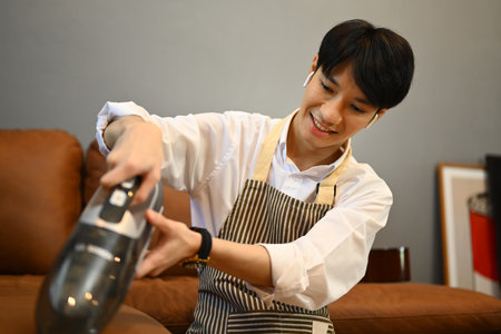 Young man cleaning sofa with cordless vacuum cleaner to removes germs and dirt. Housework concept.