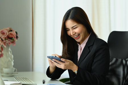 Cheerful female employee sitting at desk typing massage, communicating online on mobile phone.の写真素材