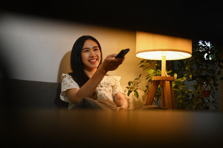 Happy young woman wearing casual clothes holding remote control, enjoying her favorite TV show at night.の写真素材