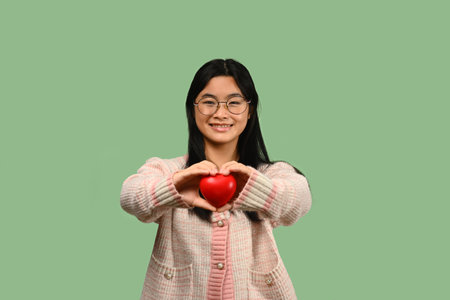 Smiling Asian teenage girl holding red heart isolated on green background. Love, valentine, gift and health symbol..の写真素材