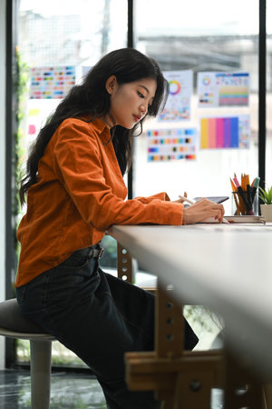 Concentrated young creative woman sitting in bright office and working on design project..の写真素材