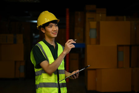 Male manager wearing safety hardhat and vest checking quantity of storage product on shelf in a large warehouse.の写真素材
