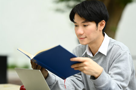 Focused asian man student reading interesting book while sitting on bench in city park. Education, lifestyle concept.の写真素材