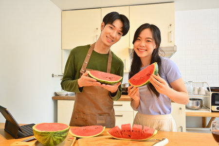 Happy young couple holding and eating slices of watermelon in home kitchen. Family love and casual lifestyle.の写真素材