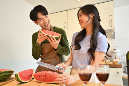 Happy married couple in aprons standing behind wooden kitchen tabletop and eating sweet watermelon.の写真素材