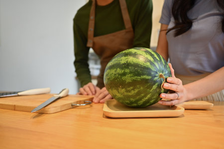 Cropped image of young couple with watermelon standing behind wooden kitchen tabletop.の写真素材