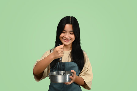 Cheerful teenage Asian girl wearing apron holding whisk and bowl isolated on green background.の写真素材