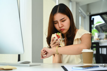 Busy asian female worker eating sandwich and checking time on watch. Fast food and daily haste and urban lifestyle concept.の写真素材