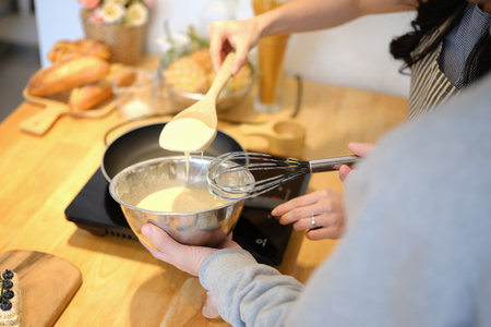 Young couple preparing breakfast, pouring batter for making pancakes with a ladle into a frying pan.