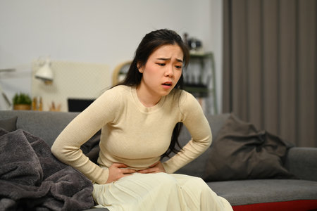 Unhealthy young woman holding belly sitting on couch at home suffering from pain, digestive...