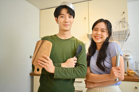 Funny young couple holding kitchenware utensils in hands and smiling at camera while cooking in the kitchen on weekend.の写真素材