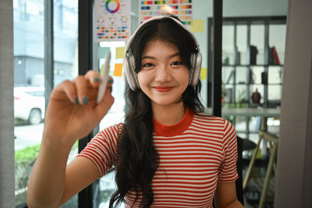 Portrait of cheerful asian woman in headphone and stripes t-shirt holding pen and pointing at camera.の写真素材