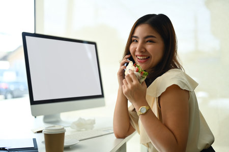 Cheerful female worker eating sandwich and talking on mobile phone at her office desk.の写真素材