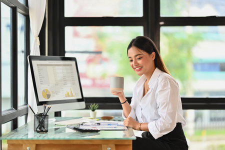 Charming female employee drinking coffee and reading sales reports at her workplace.の写真素材