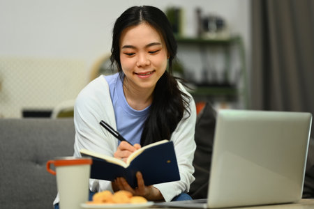 Beautiful young asian woman using laptop and making notes on notebook.