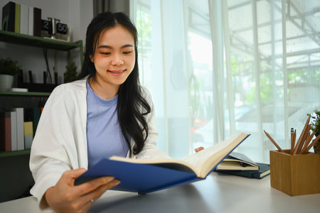 Happy young Asian woman in casual clothes reading book on table in living room. Education and leisure activity concept.の写真素材