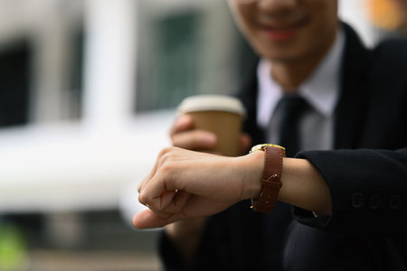 Businessman holding take away coffee cup and checking time on wristwatch for appointment or...