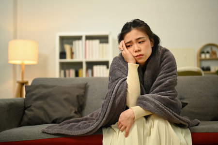 Sick woman suffering from strong headache or migraine while covered with a blanket sitting on sofa.
