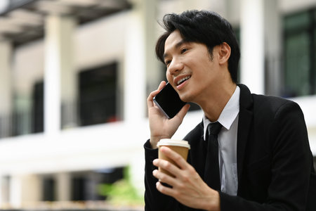 Positive asian businessman having pleasant phone conversation on mobile phone sitting outside office building.の写真素材