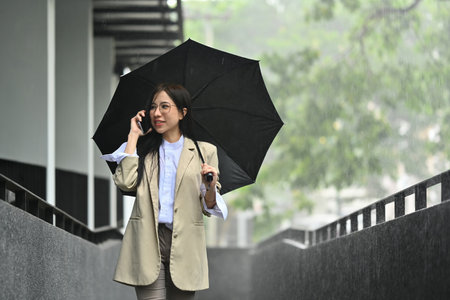Gorgeous millennial businesswoman talking on mobile phone under umbrella while walking in the city...