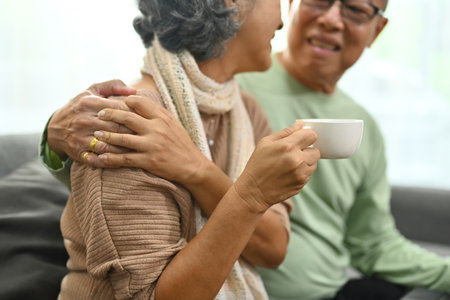 Carefree middle aged old husband embracing wife, mature woman holding cup of tea, relaxing on sofa.の写真素材
