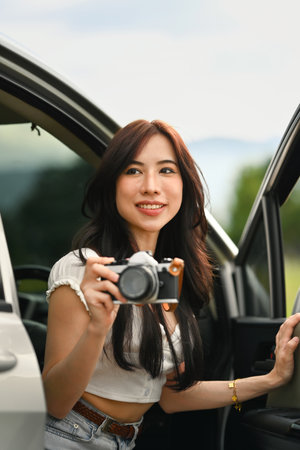 Charming millennial woman sitting in car with open door taking photos with vintage retro cameras during weekend road trip.の写真素材